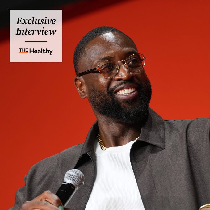 A smiling man with a beard and glasses holds a microphone, wearing a gray jacket and white shirt, in front of a red background. Text reads: 